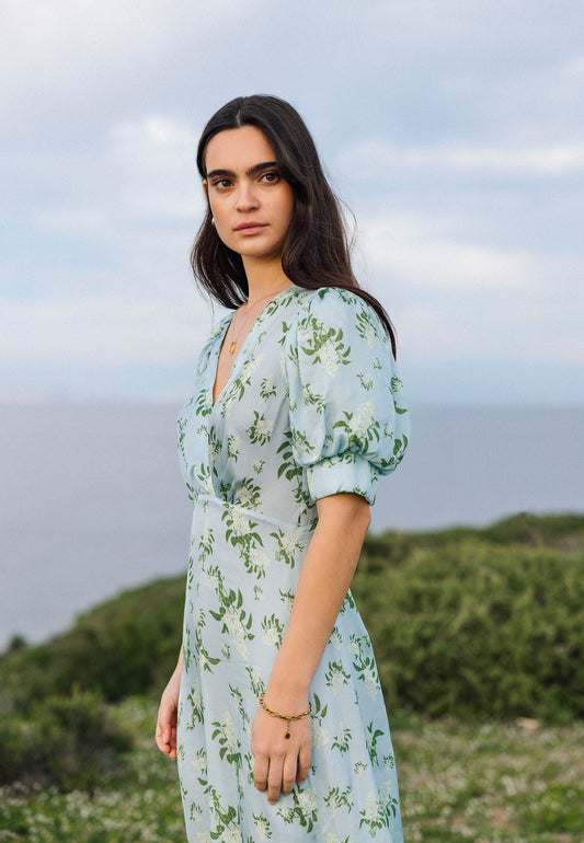 A woman in a blue TILDA dress with a floral pattern and short buff sleeves, standing against the sea and sky, looks seriously towards the camera.