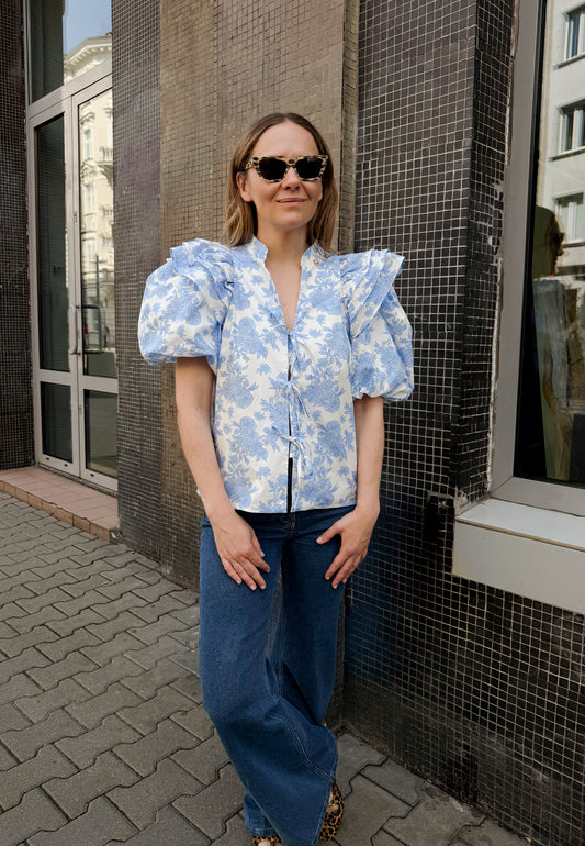 The model standing in front of the building wearing an ALIANNA blouse in a floral print in blue with front ties and ATLANTA pants.