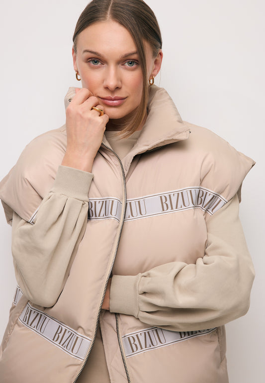 Close-up of the model with her hand near her face, wearing a beige AYASE down vest with branded tape.