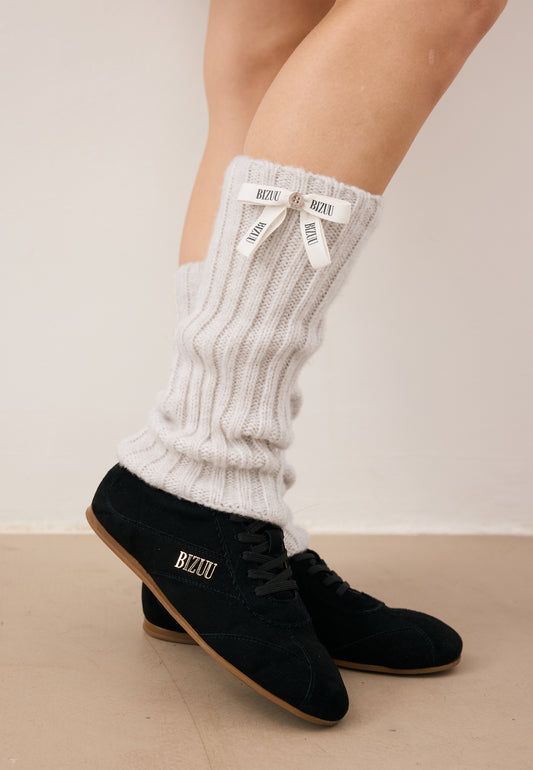Close-up of the model’s legs, with one foot resting on her toes. She is wearing beige **CHAFI** leg warmers and LEOLA shoes.