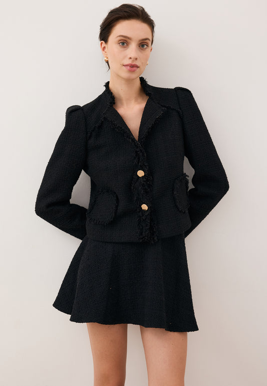 Model leaning against a wall with hands behind her back, wearing the WENDYS mini skirt and the black DITAS blazer with branded buttons.
