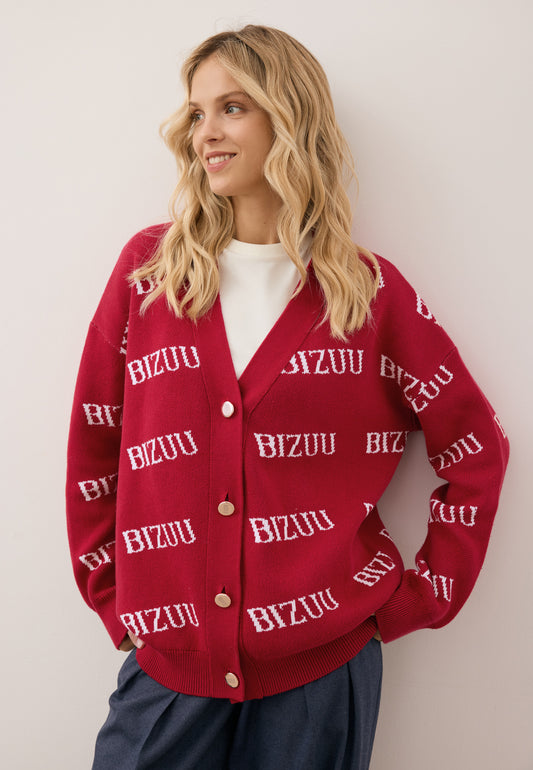 Close-up of the model leaning against a wall wearing the burgundy KYLE cardigan with a logo pattern, fastened with golden buttons.