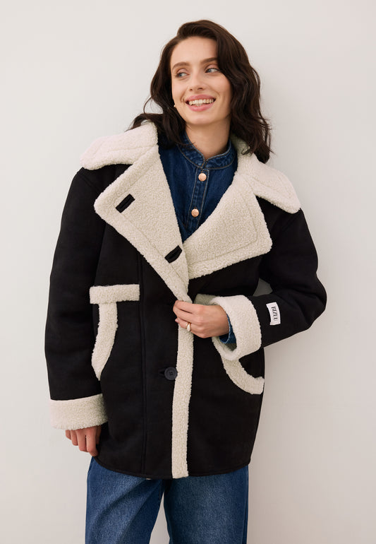 Smiling model fastening a button on the black LAFARA shearling jacket with a decorative logo on the sleeve.