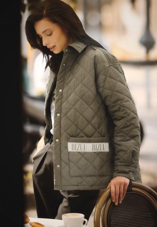 The model is looking down, leaning on a chair with one hand. She is wearing a khaki MARIN quilted jacket.