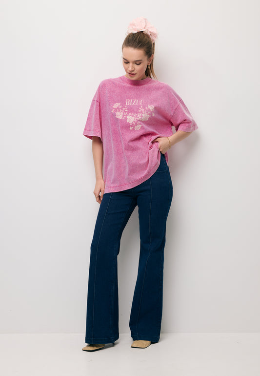 The model against a white wall with one hand resting on her hip, wearing a pink oversized PAPLYS T-shirt and navy BELINNA flared jeans.