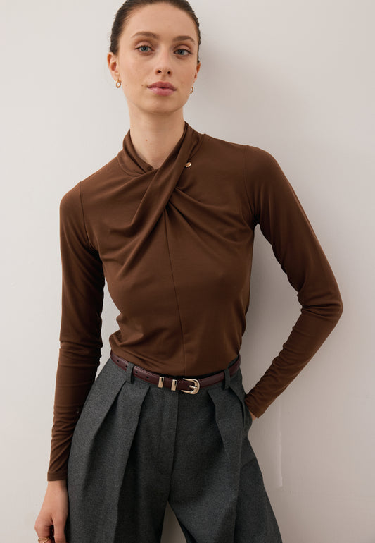 Close-up of the model with one hand behind her back wearing the chocolate brown PINOWA long-sleeve with a twist detail and wide-leg BANOS trousers.