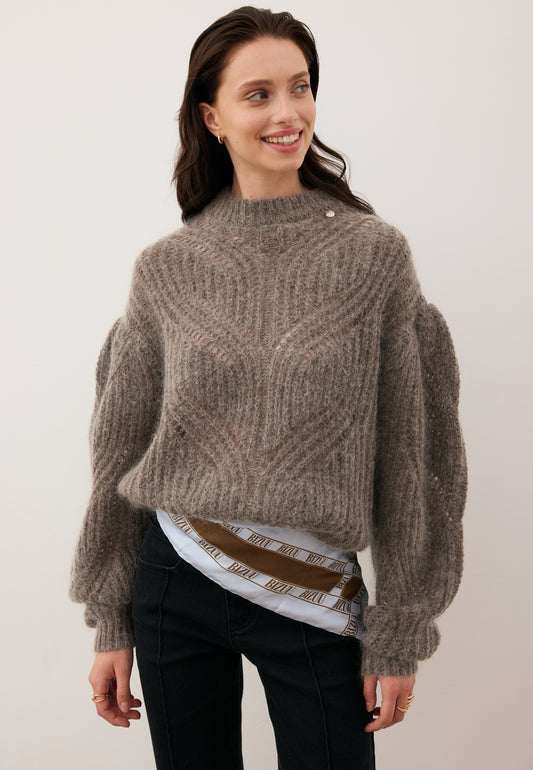 Smiling model looking to the side wearing the beige ROSSO openwork sweater with a light blue ARIA scarf tied around her hips.