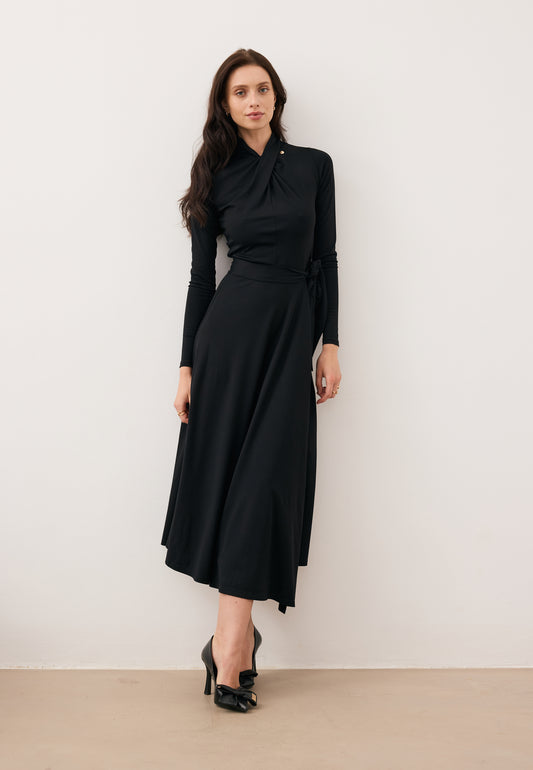 Model leaning against a wall wearing the black midi ROTTA dress with a decorative twist at the neckline.