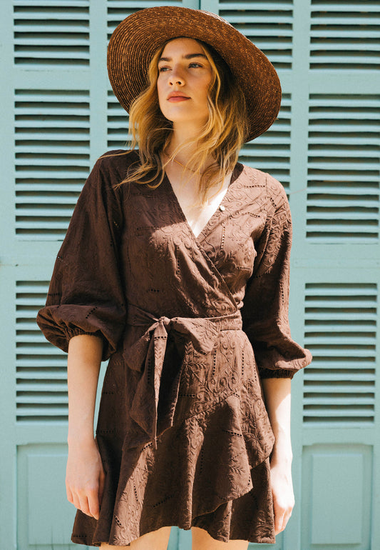 Close-up on the model in a dark brown dress with English embroidery, envelope neckline and buff sleeves AMBROSIA; a straw hat on her head, turquoise shutters in the background.