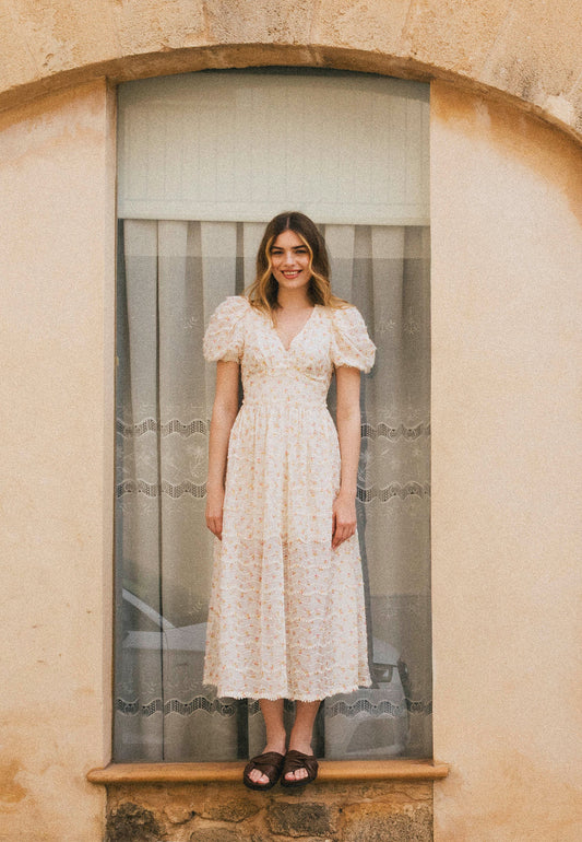 A smiling woman in a romantic midi dress in pastel flowers with FIFFI buffets, stands on the window sill with a curtain.