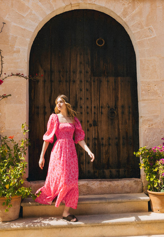 A woman in a pink floral LUMIA dress with buff sleeves stands in front of a large wooden door in a stone wall, surrounded by potted flowers.