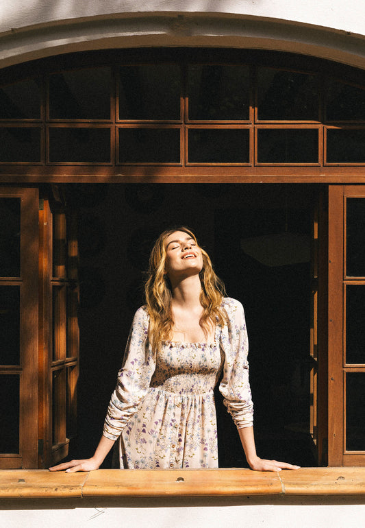 A model wearing a long PATRYA dress with a delicate floral pattern leans out of a wooden window, her face facing the sun with her eyes closed, in the light of the morning glow.