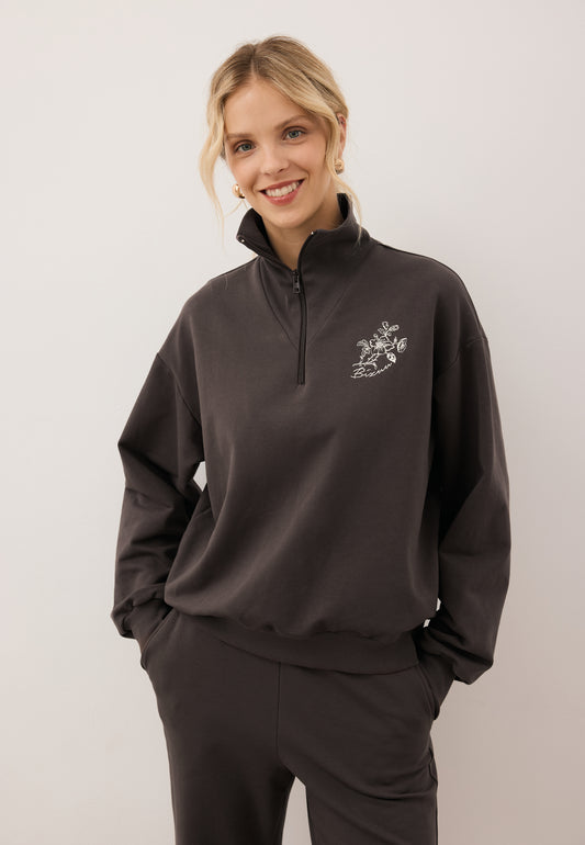 Smiling model looking into the camera with hands in the pockets of the ZEIA trousers. Close-up of the brown TANSY sweatshirt with front logo appliqué.