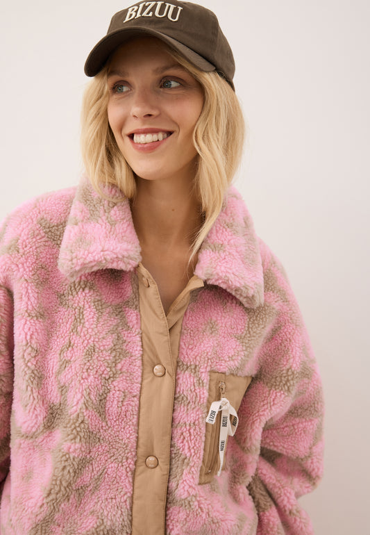 Close-up of the smiling model wearing the floral TEDDY fleece jacket with decorative branded bow and the brown ZANE cap.