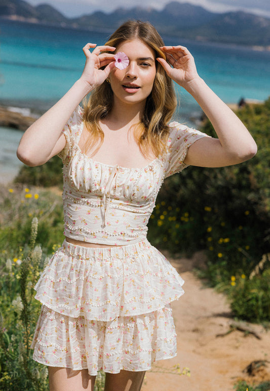 A smiling woman in a romantic set with VANNES and COKO ruffles poses on a path among wildflowers, holding a pink flower to her eye. In the background is a blue lagoon and a mountainous landscape.