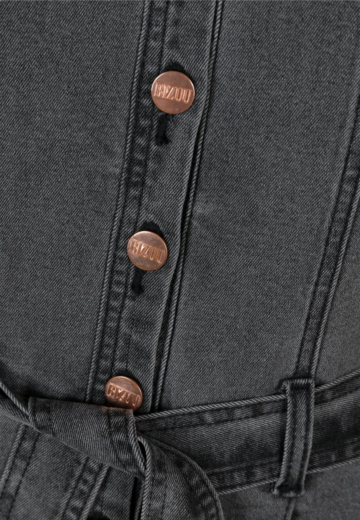 Close-up of a dark gray denim jumpsuit with four copper buttons bearing the inscription “BIZUU,” a visible belt loop, and a fragment of a tied belt.