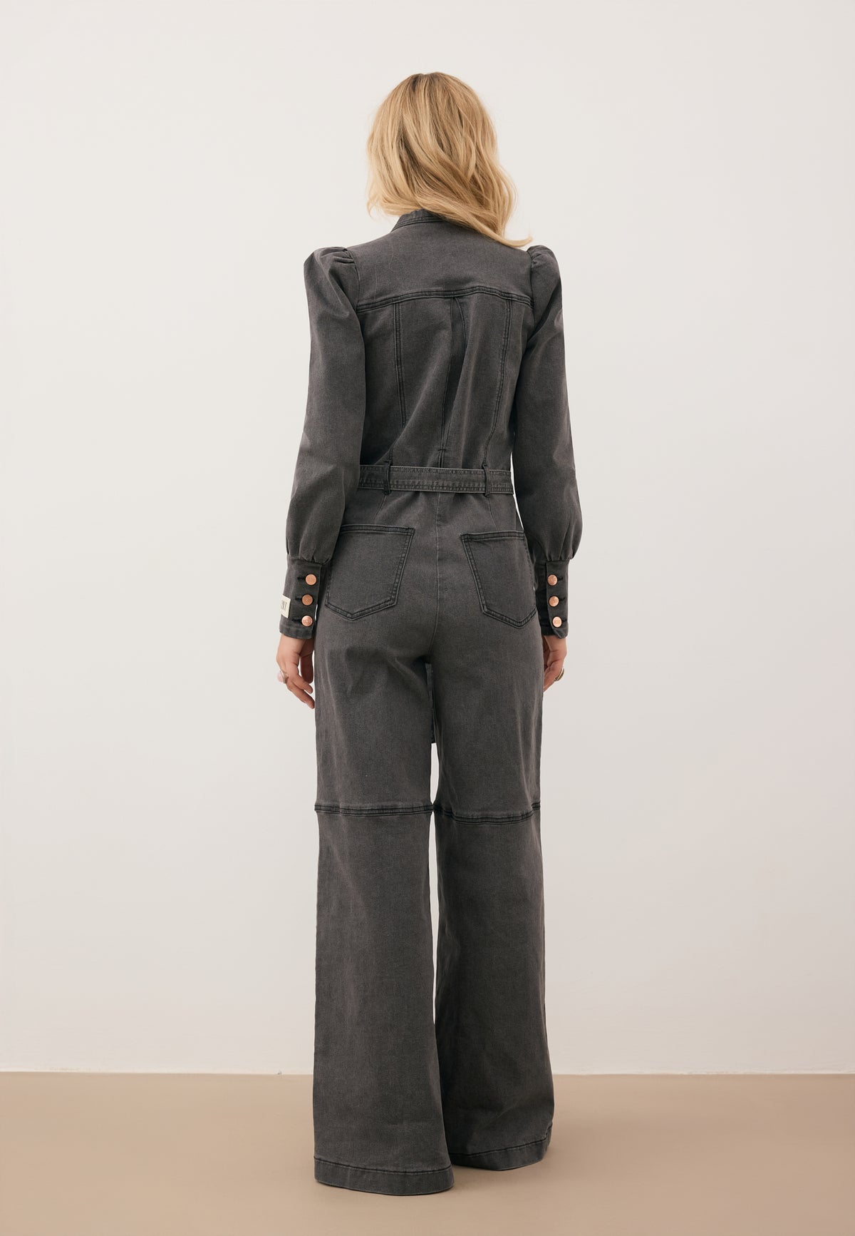 A model standing with her back to the camera, wearing a gray MENDINA denim jumpsuit tied at the waist.