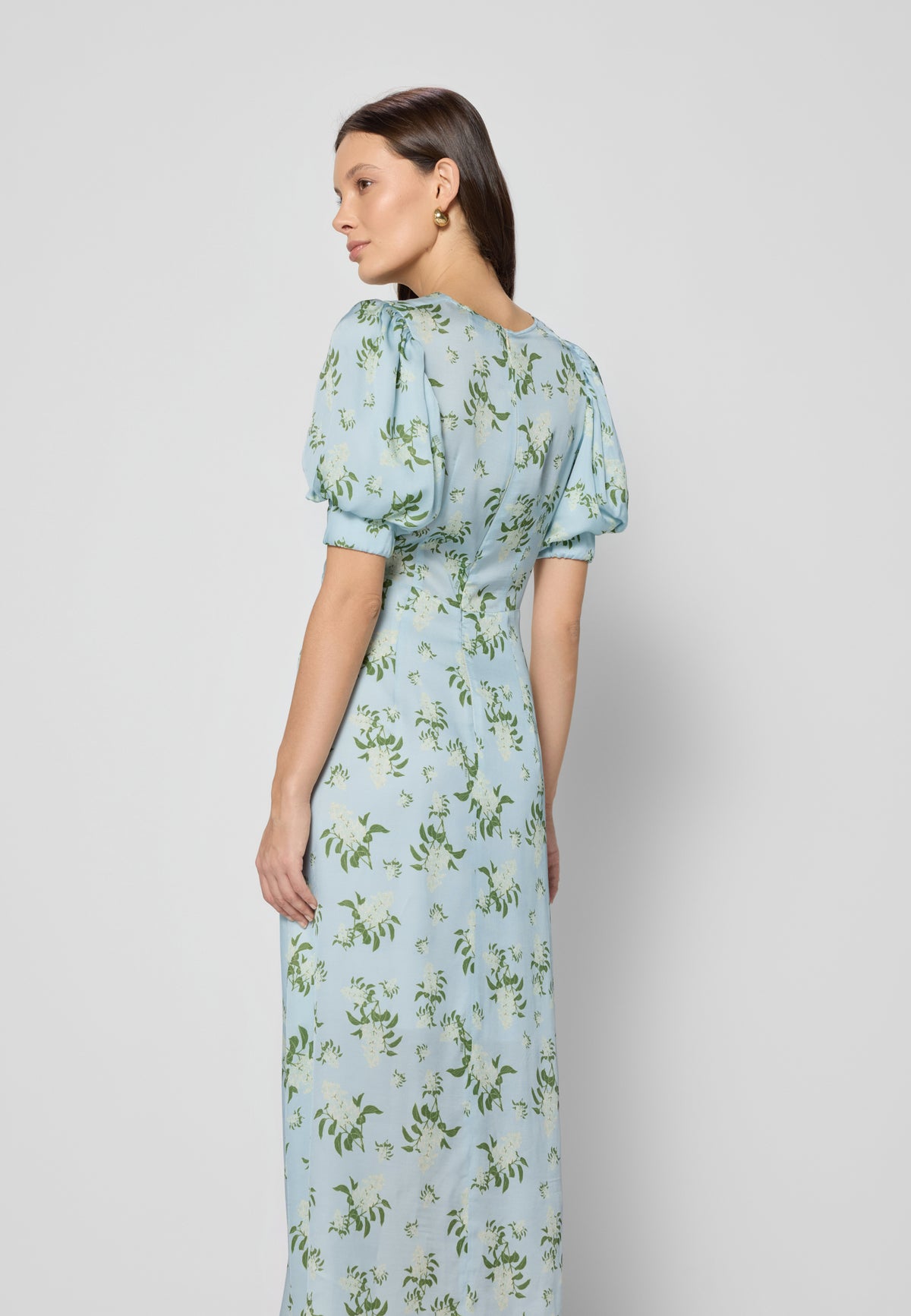 A woman in a blue back dress, showing the back of the TILDA dress and buff sleeves, against a gray studio background.