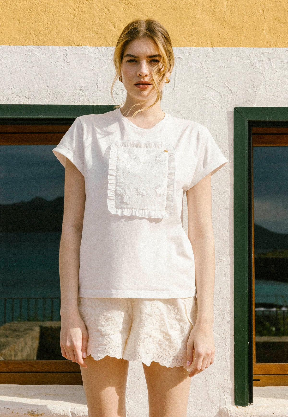 Close-up of the model standing against a colorful wall and windows in a cream MIRADO T-shirt and NILDA lace shorts.