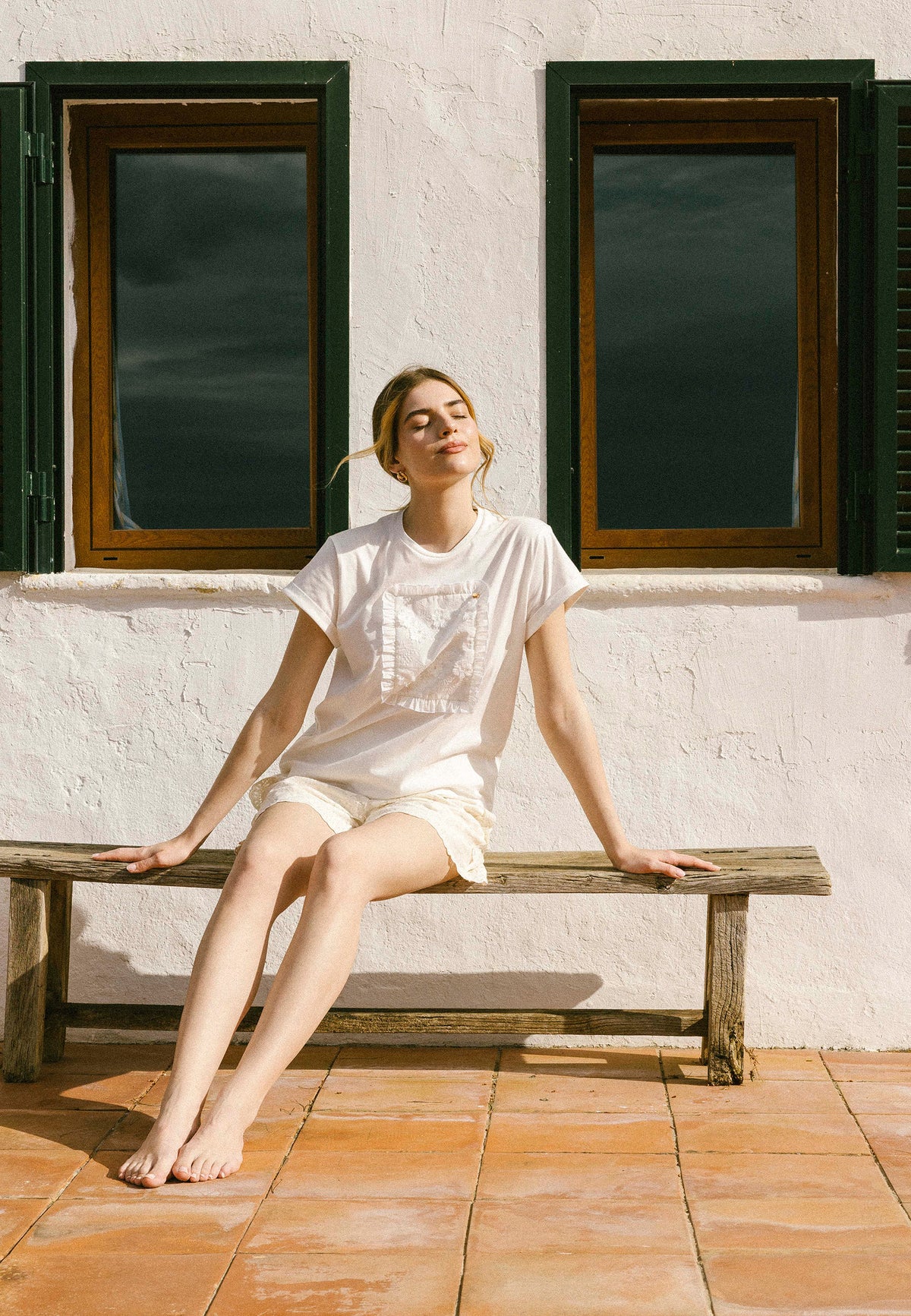 Model sitting on a wooden bench against a background of a colorful wall and windows in a cream MIRADO T-shirt and NILDA lace shorts.