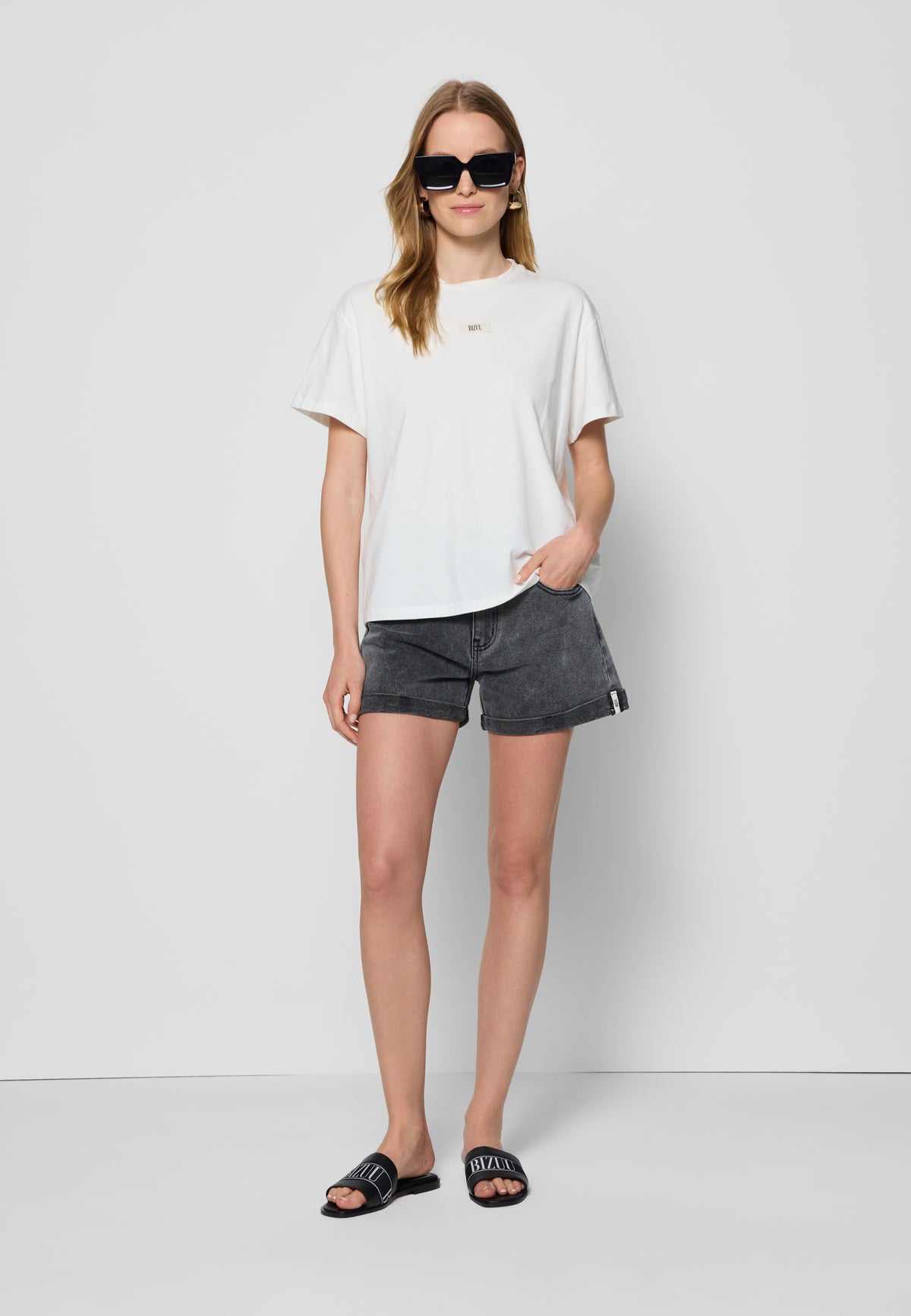 The model on the white background wearing a cream TISHALLE T-shirt, MENORCA denim shorts and TANNY sunglasses.