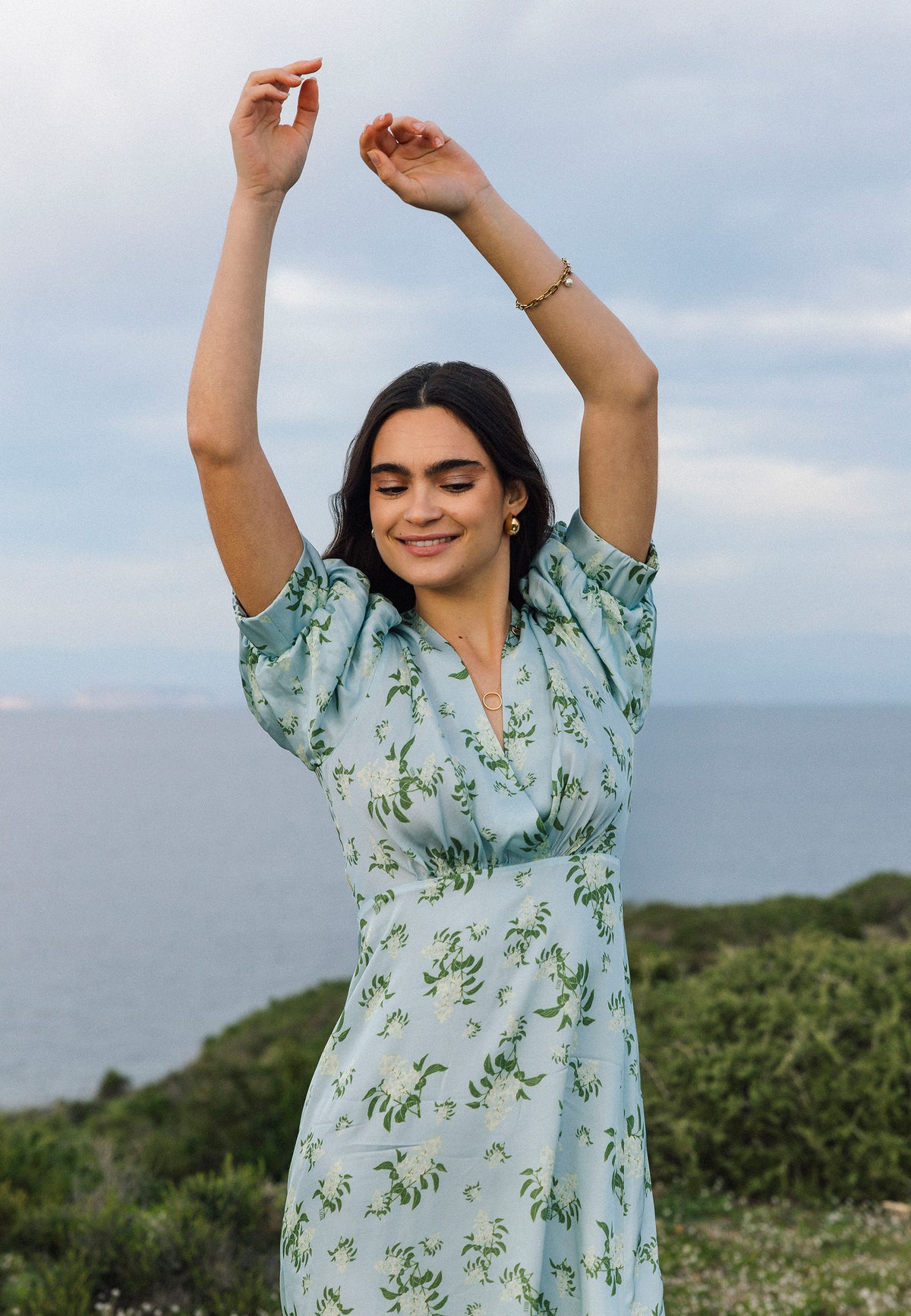 A smiling TILDA woman in the same dress raises her hands in a dancing pose against the sea.