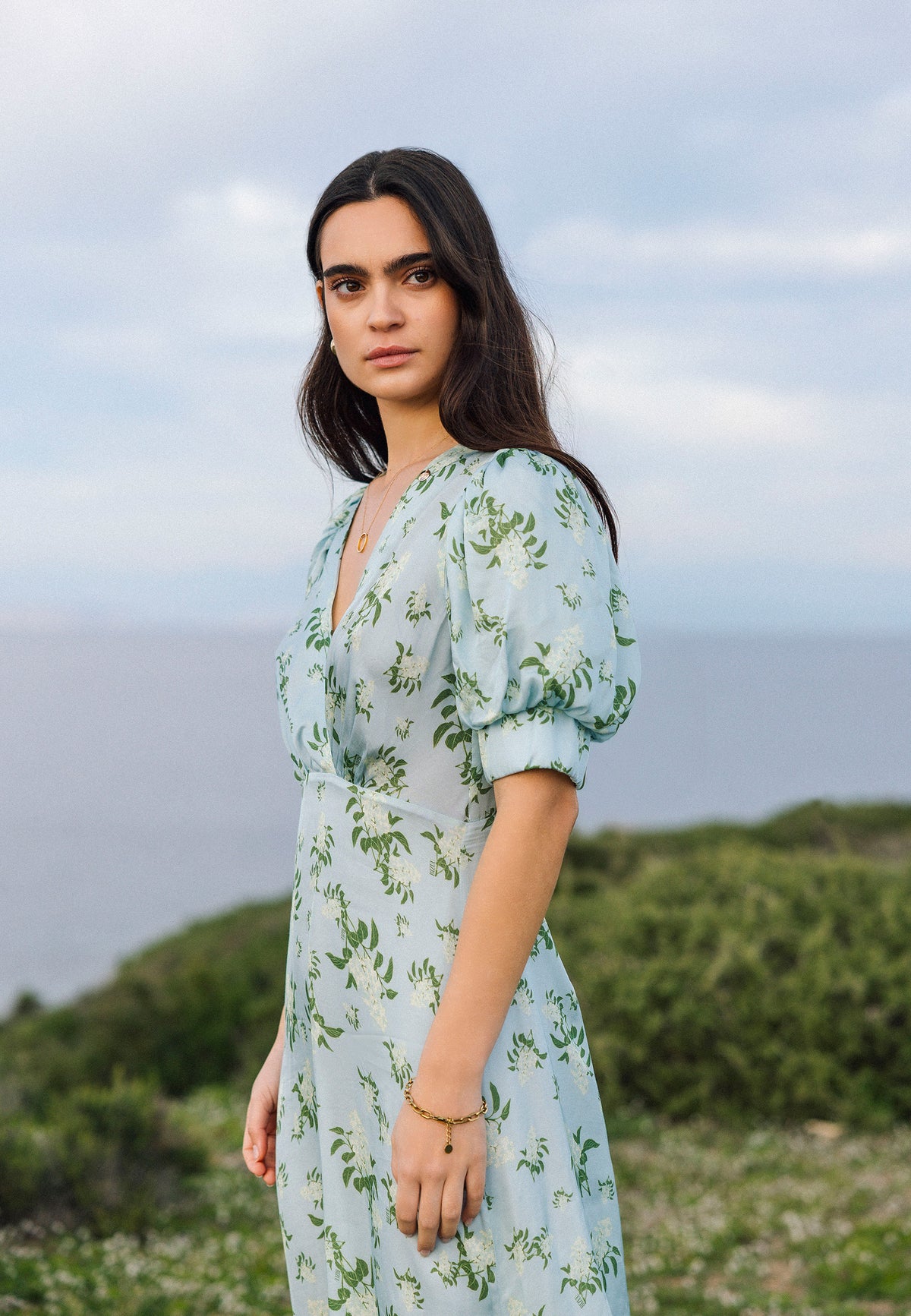 A woman in a blue TILDA dress with a floral pattern and short buff sleeves, standing against the sea and sky, looks seriously towards the camera.