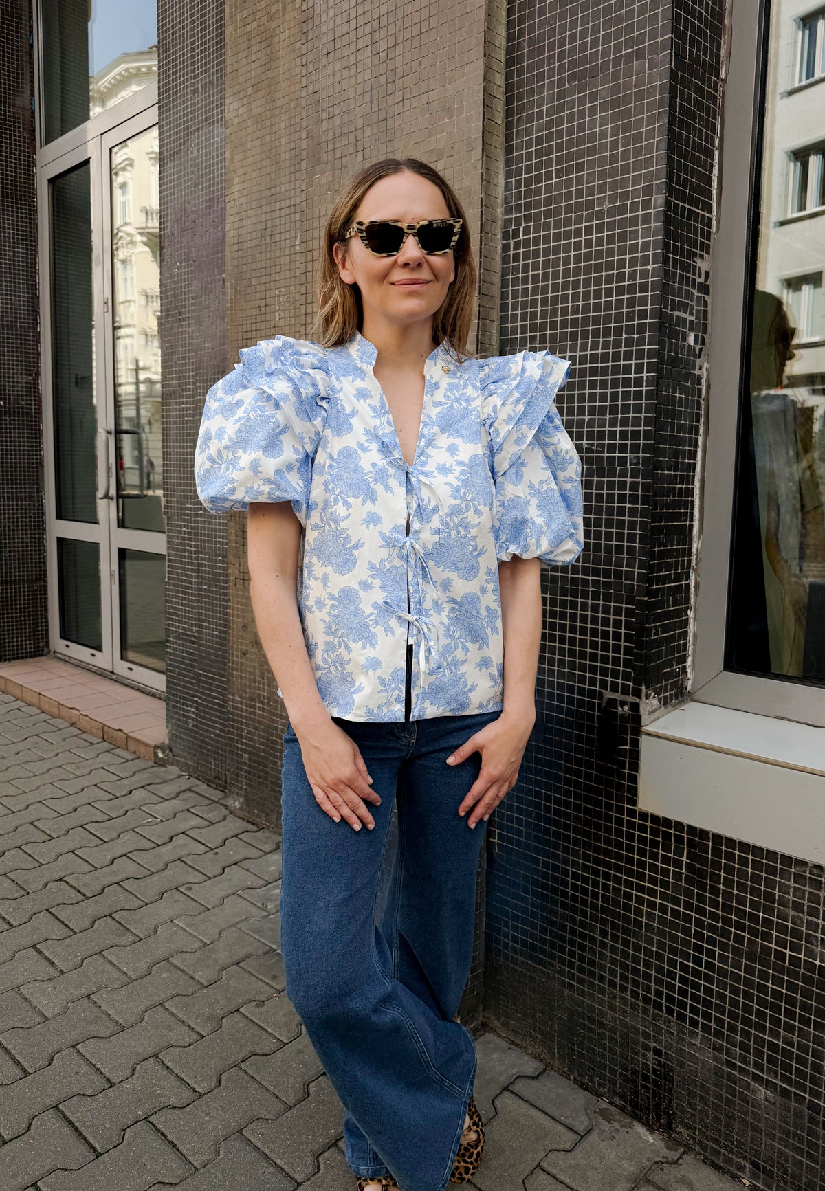 The model standing in front of the building wearing an ALIANNA blouse in a floral print in blue with front ties and ATLANTA pants.