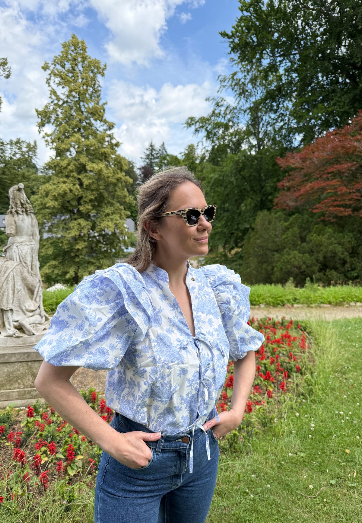 The model standing in the garden with her hands stuck in her pockets. She is wearing an ALIANNA cotton blouse in a blue floral print.