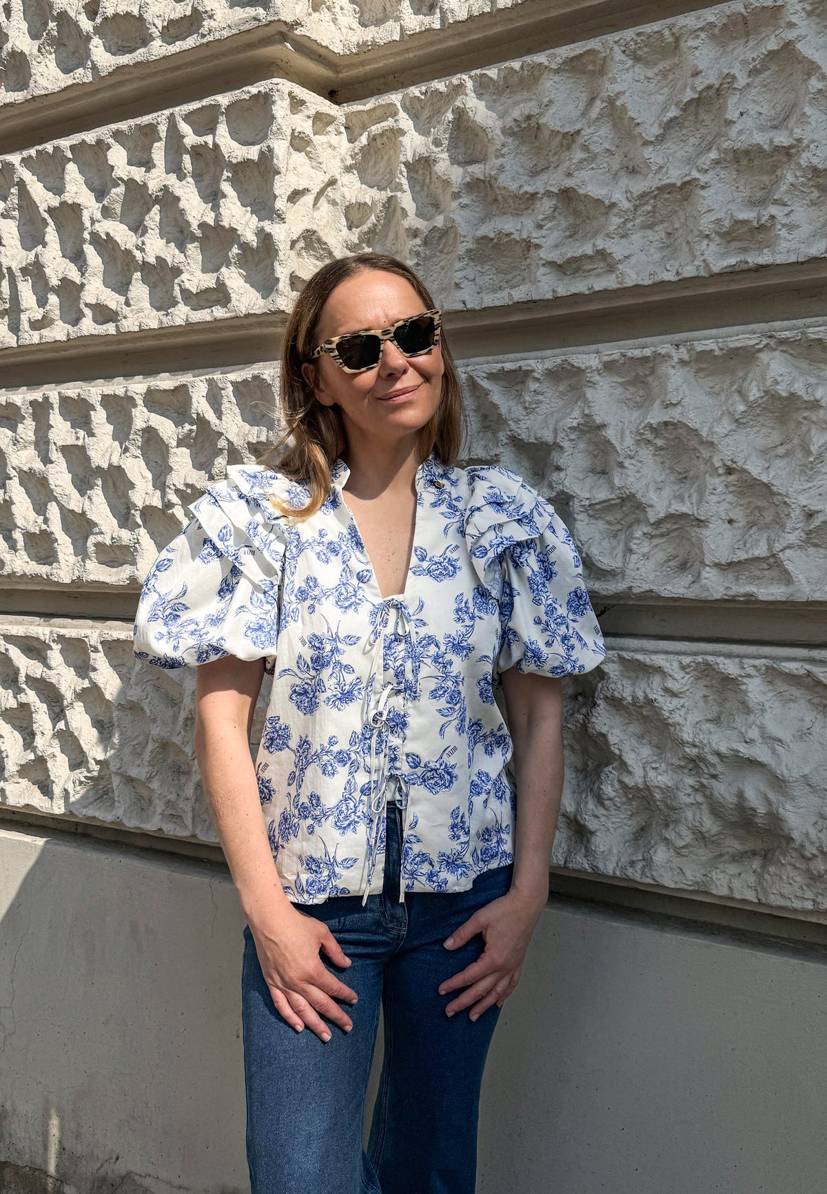 Model standing against the wall of the building wearing ALIANNA cotton blouse with front ties and buff sleeves.