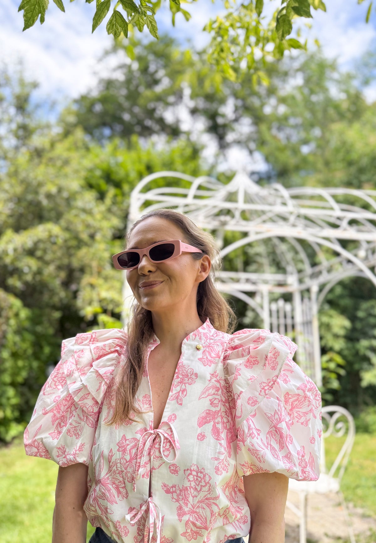 A close-up of the model standing in the garden, wearing an ALIANNA blouse tied at the front in a floral print.
