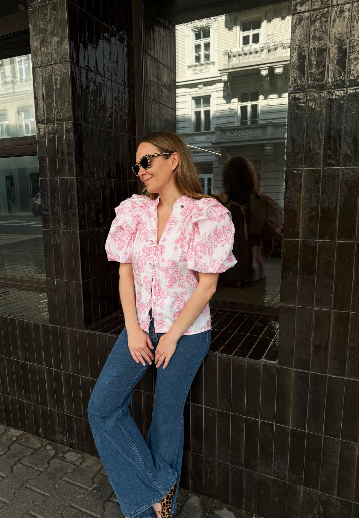 The model is leaning against a windowsill with her legs crossed. She is wearing an ALIANNA cotton blouse with ties at the front in a floral white and pink print.