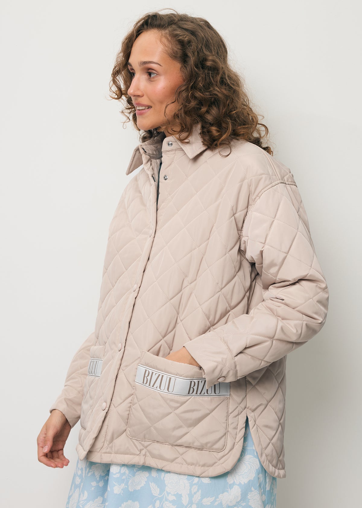 A model standing sideways, wearing the beige MARIN jacket with quilting and a logo on the pocket.