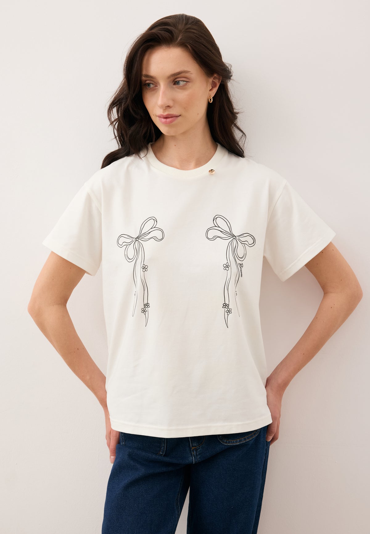 Model leaning against a wall with hands on hips, styled in the cream BOW T-shirt with bow illustration and LEYA trousers.