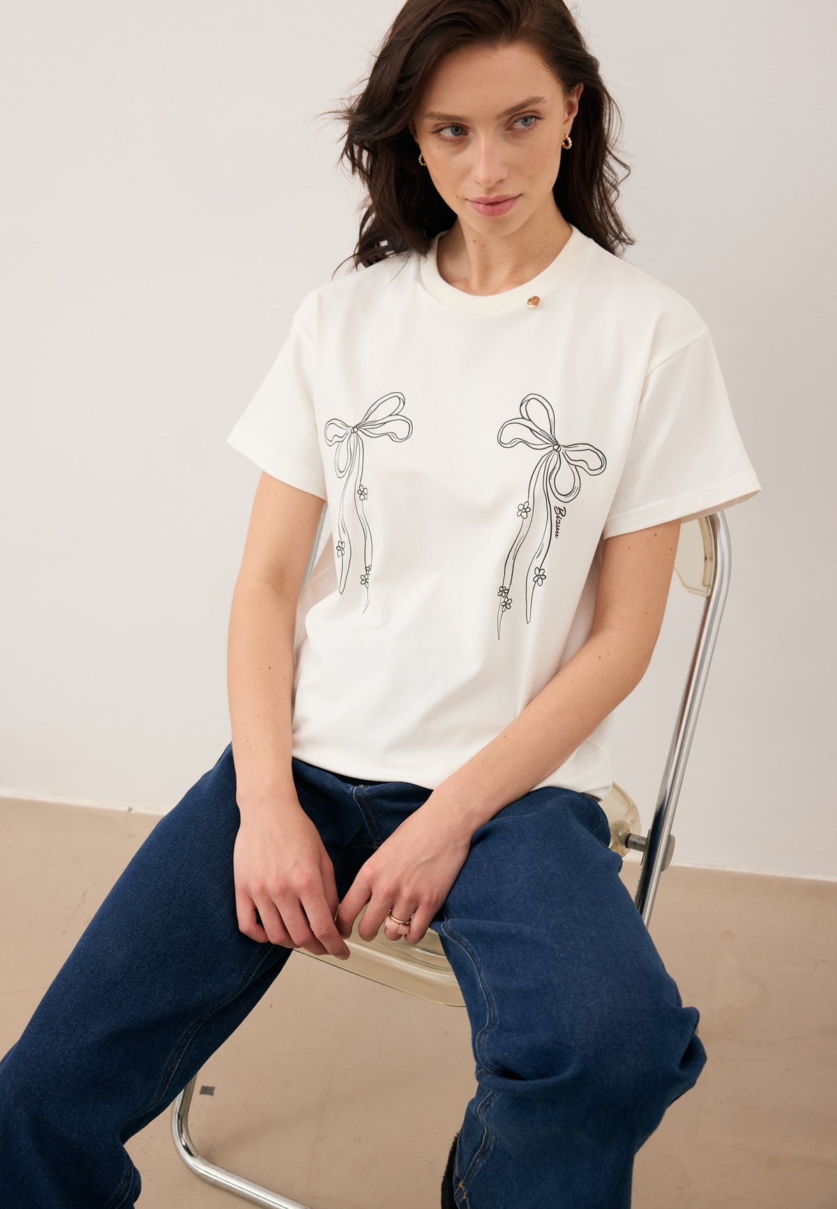 Model sitting on a chair, dressed in the cream BOW T-shirt with bow print and logo detail, paired with navy LEYA jeans.