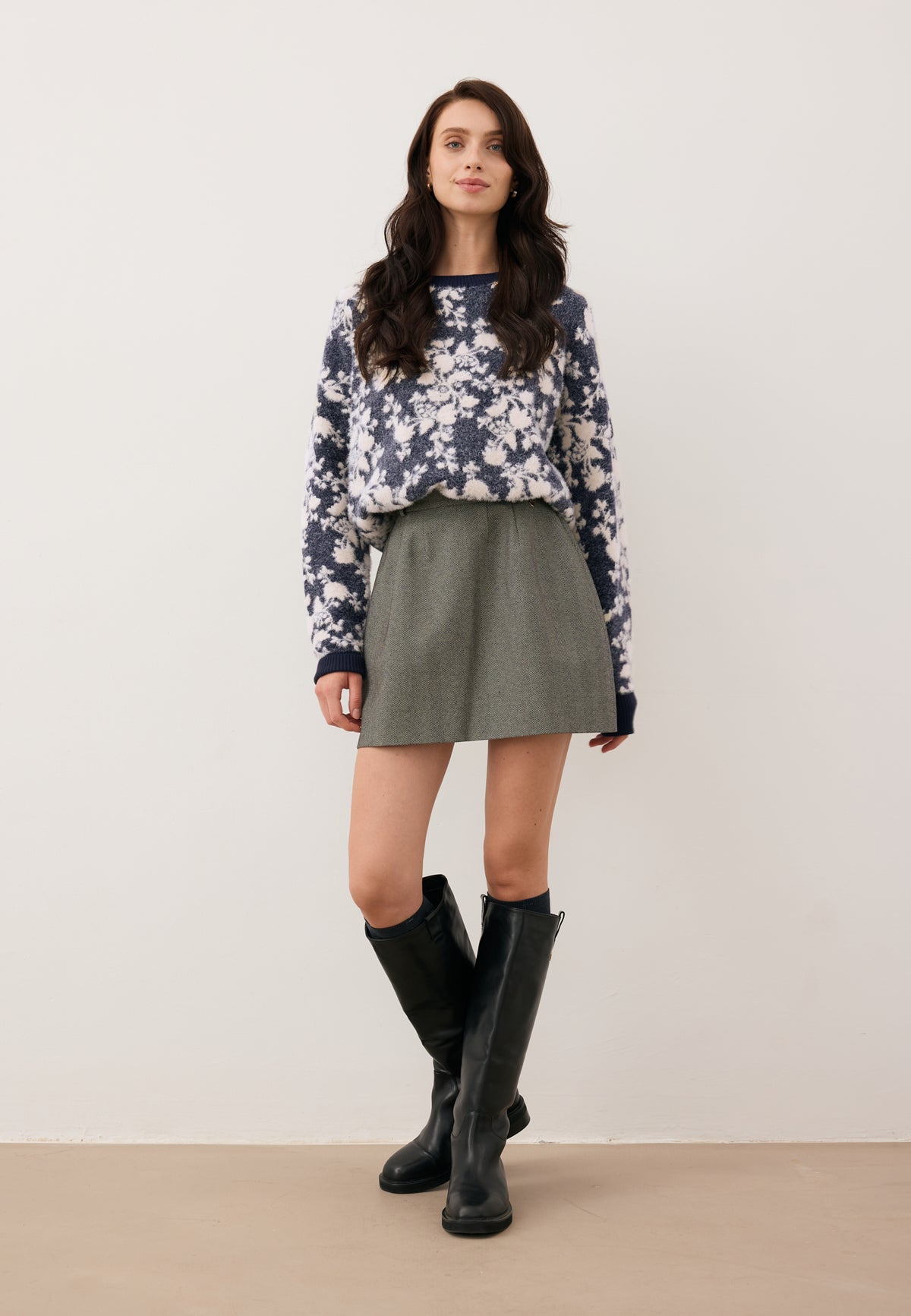 Model stepping forward against a white wall, wearing the floral CANELA sweater, BREDA herringbone mini skirt, and black CALYX boots.