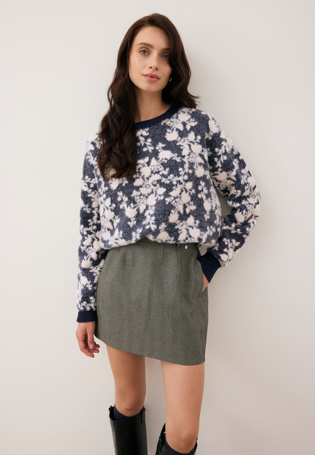 Model leaning against a wall dressed in the floral CANELA sweater and the black BREDA mini skirt.