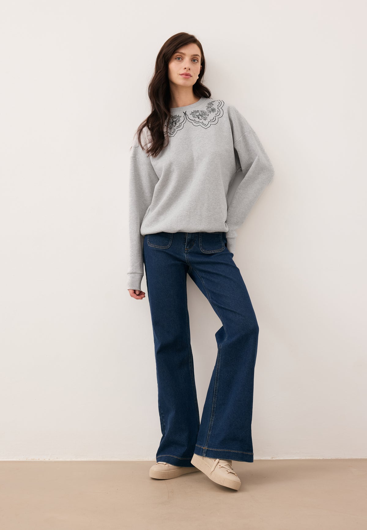 Model leaning against a wall with one leg bent, dressed in the gray COLLIER sweatshirt with decorative collar print and navy DELPHI jeans.