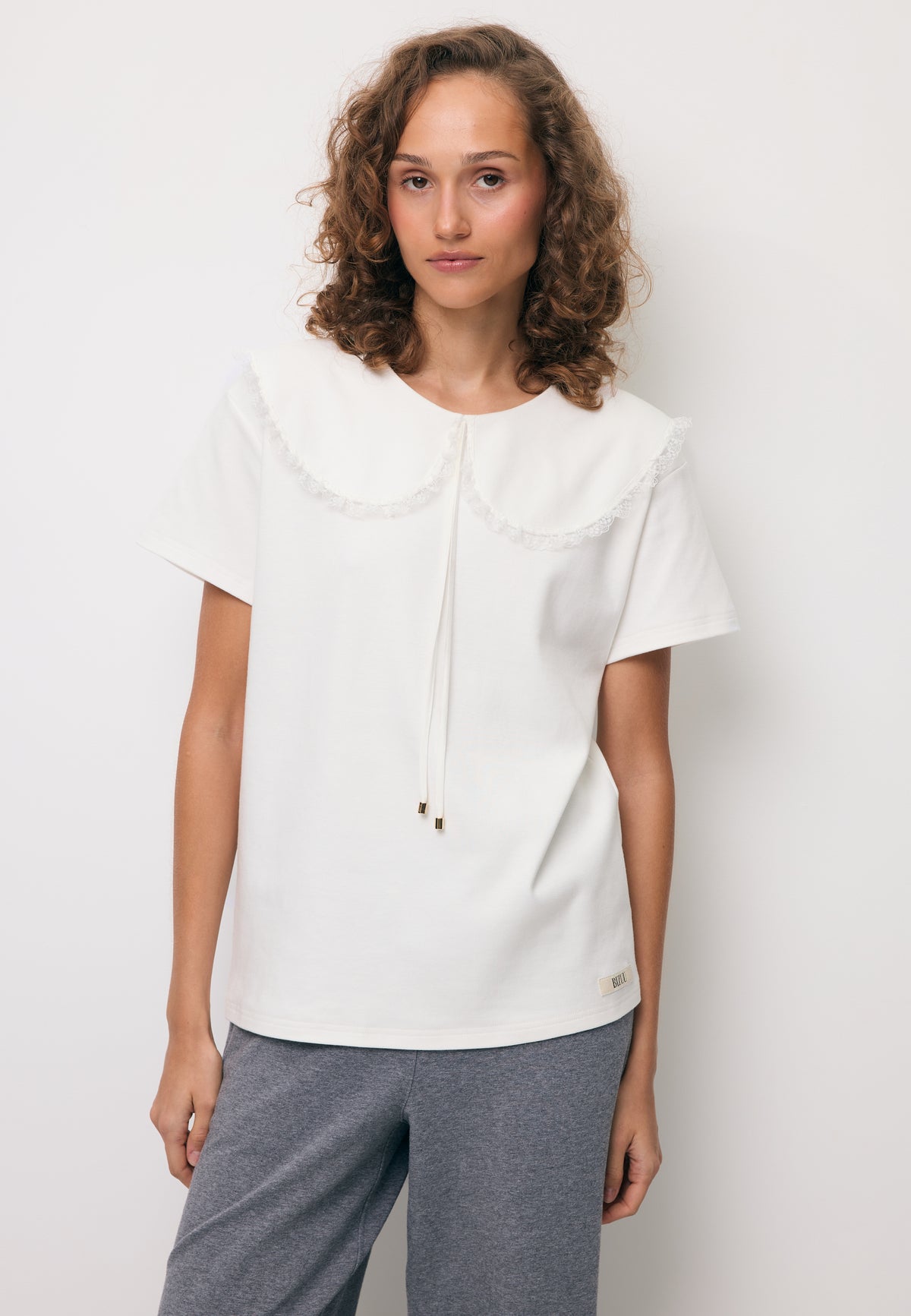 Close-up of a model wearing a cream CUTIE collared shirt and grey BAYLE frilled trousers.