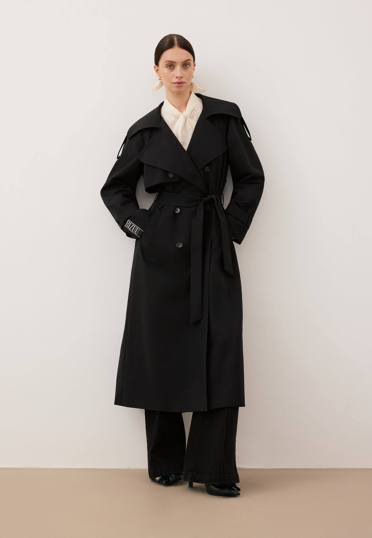 A model leaning against a white wall with her hands behind her back, wearing a black RACHELLA coat tied at the waist and a DEFIORI bodysuit.