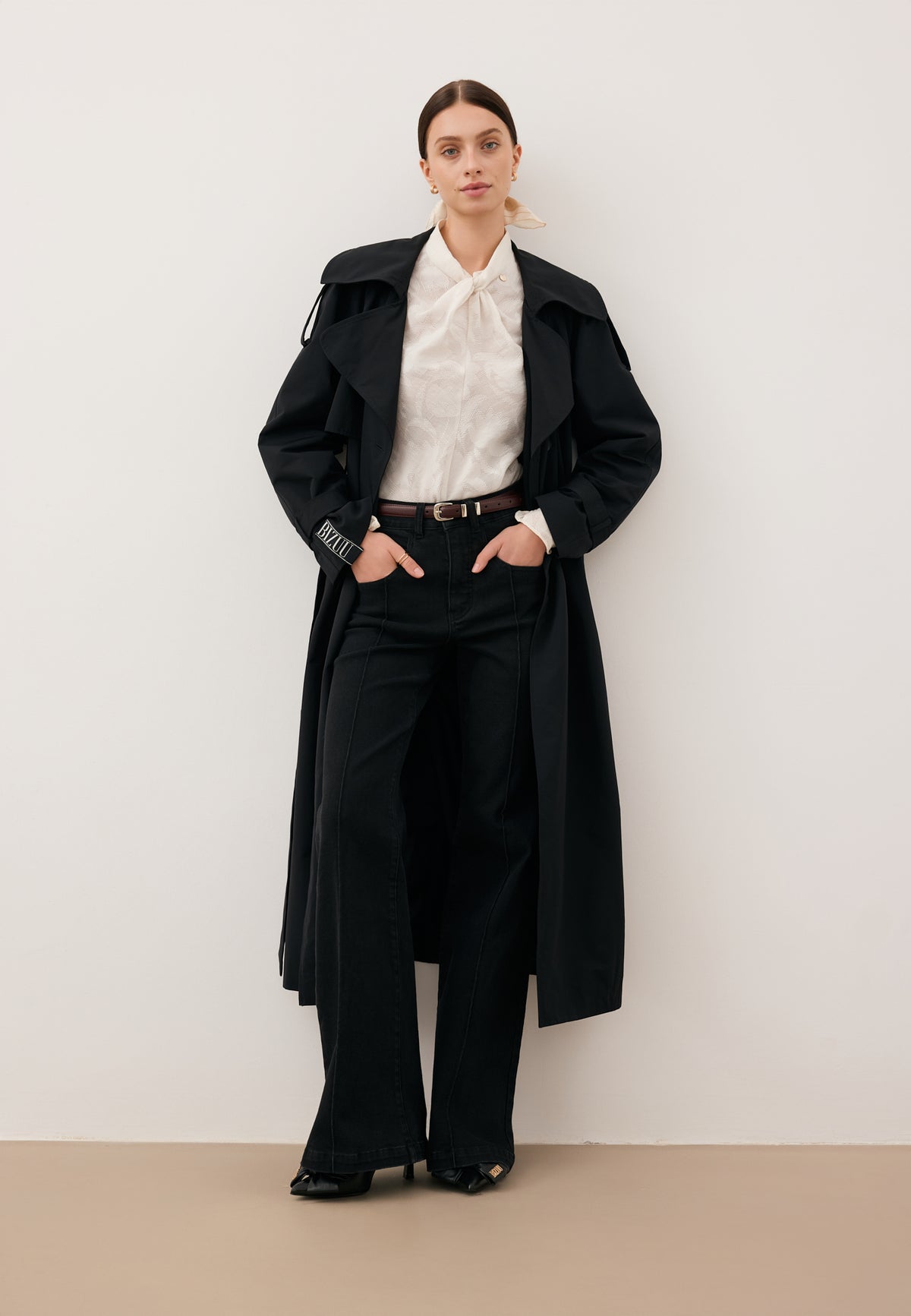A model leaning against a white wall with her hands in the pockets of her black BELINNA pants and wearing an unbuttoned black RACHELLA coat.