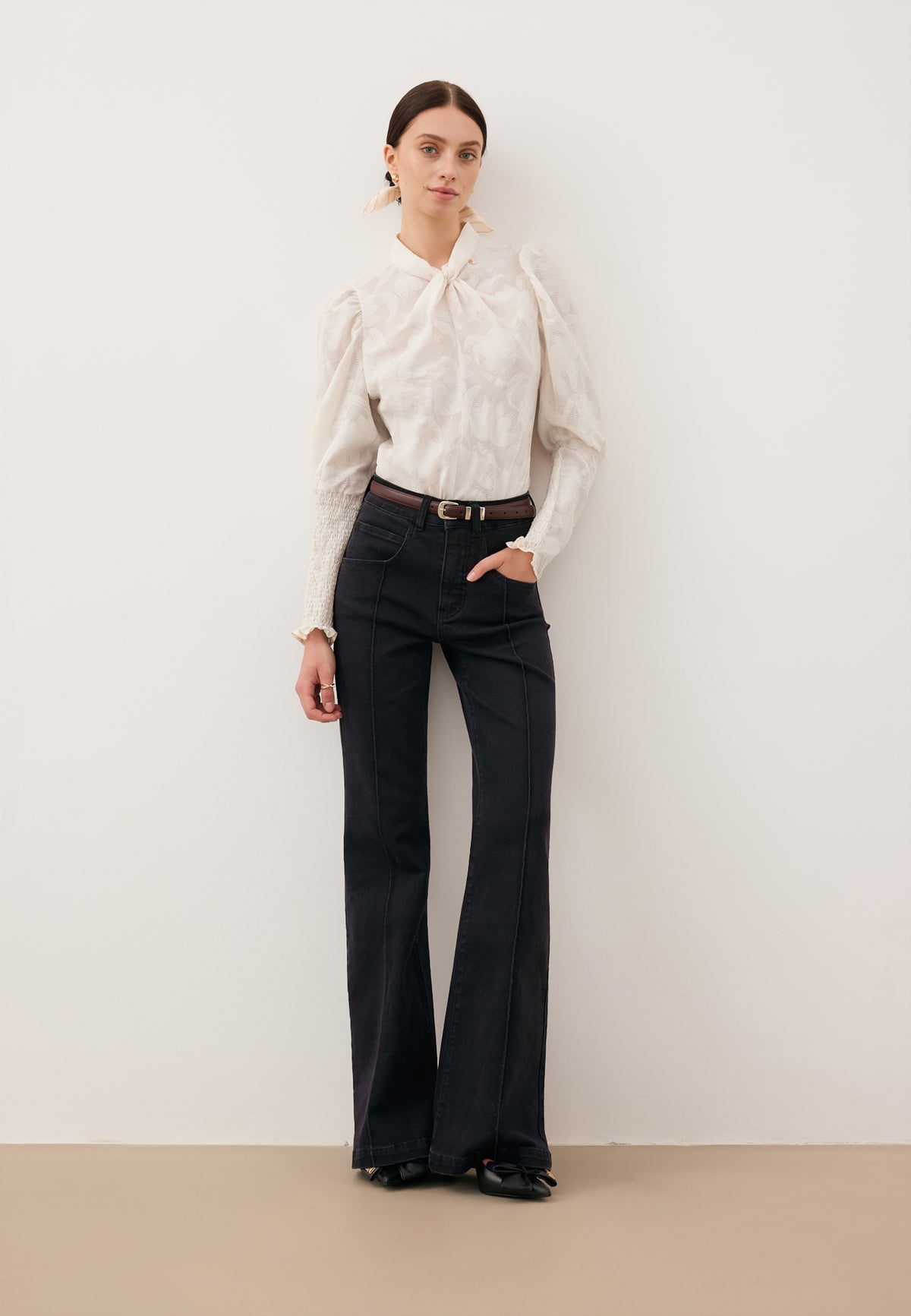 A model leaning against a white wall, wearing black BELINNA flare jeans and a cream DEFIORI bodysuit.
