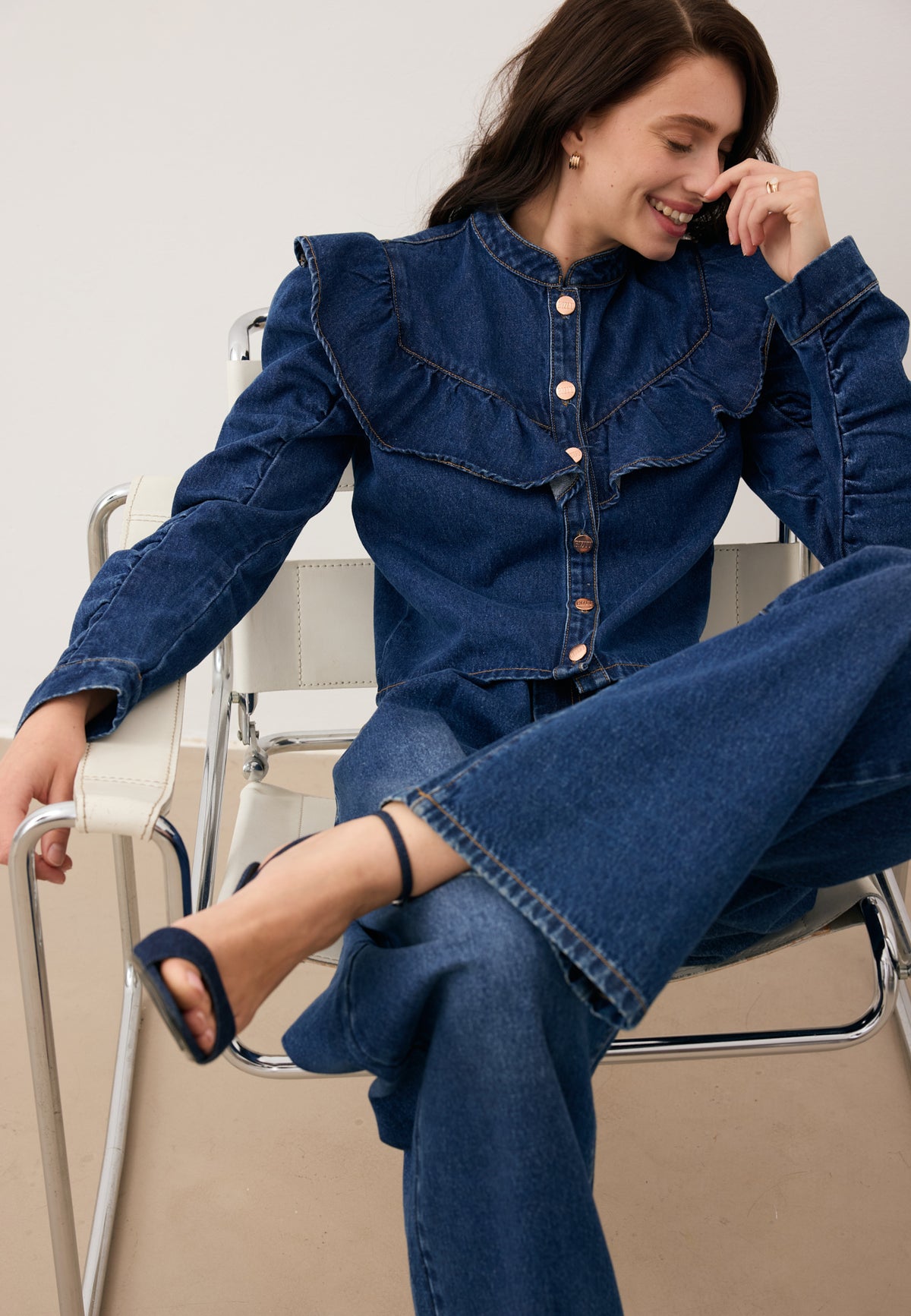 Model sitting on a chair, laughing with one leg crossed over the other, dressed in the navy denim set – DILETTA shirt and LEYLA jeans.