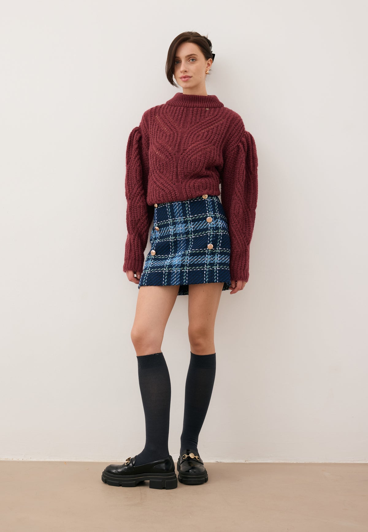 Model leaning against a wall, dressed in the DISSA navy check mini skirt and the burgundy ROSSO sweater.