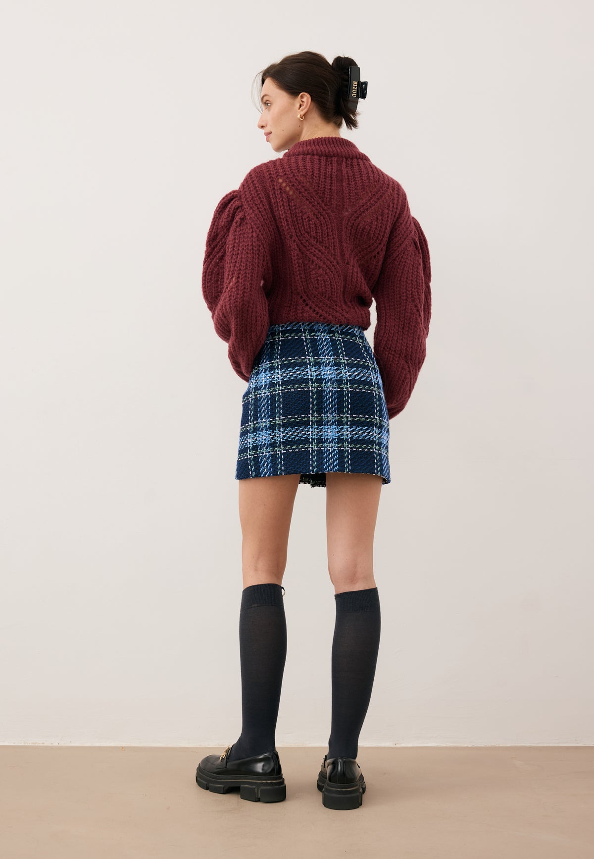 Model standing with her back to the camera, dressed in the burgundy ROSSO sweater, DISSA navy mini skirt in a check pattern, SCHOOLI knee-high socks, and LESOTHO shoes.