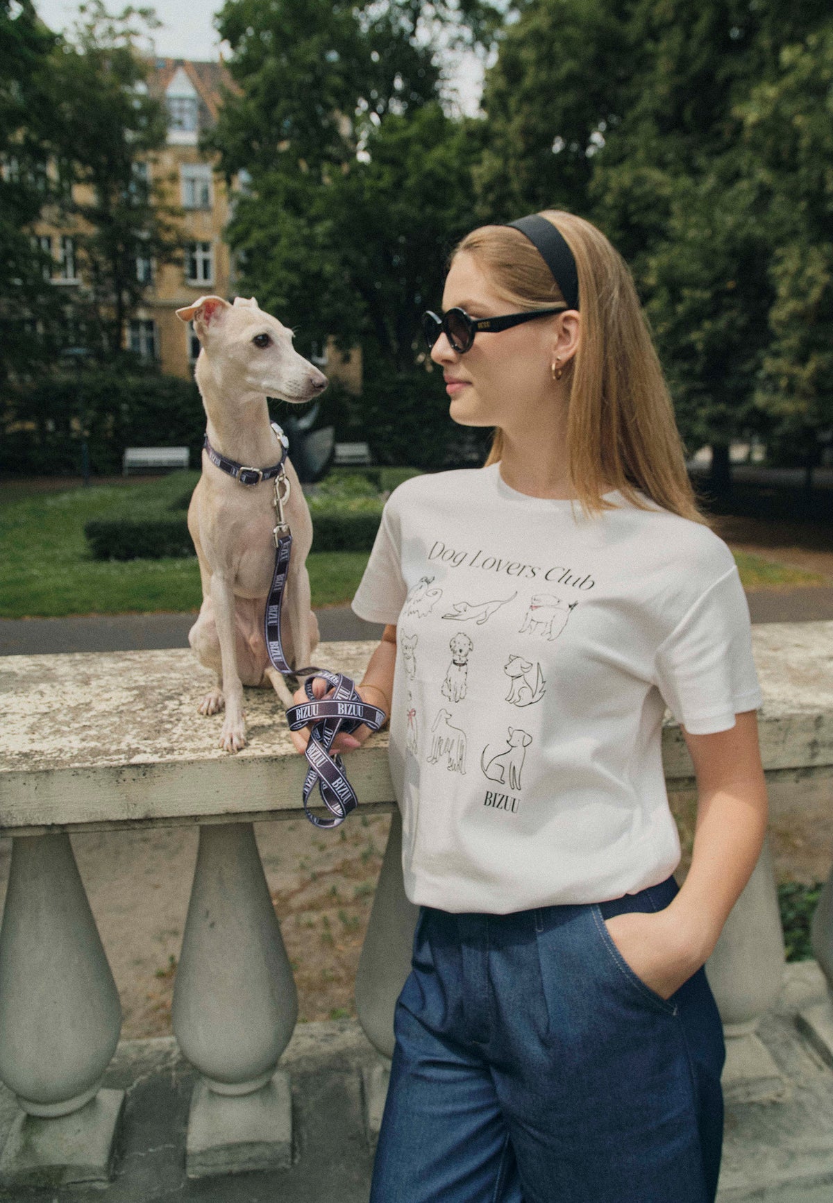 A model leaning against a wall with a dog sitting on it. She is wearing a white T-shirt with dog drawings and the inscription Dog Lovers Club. She is holding a leash with the Bizuu logo in her hand.