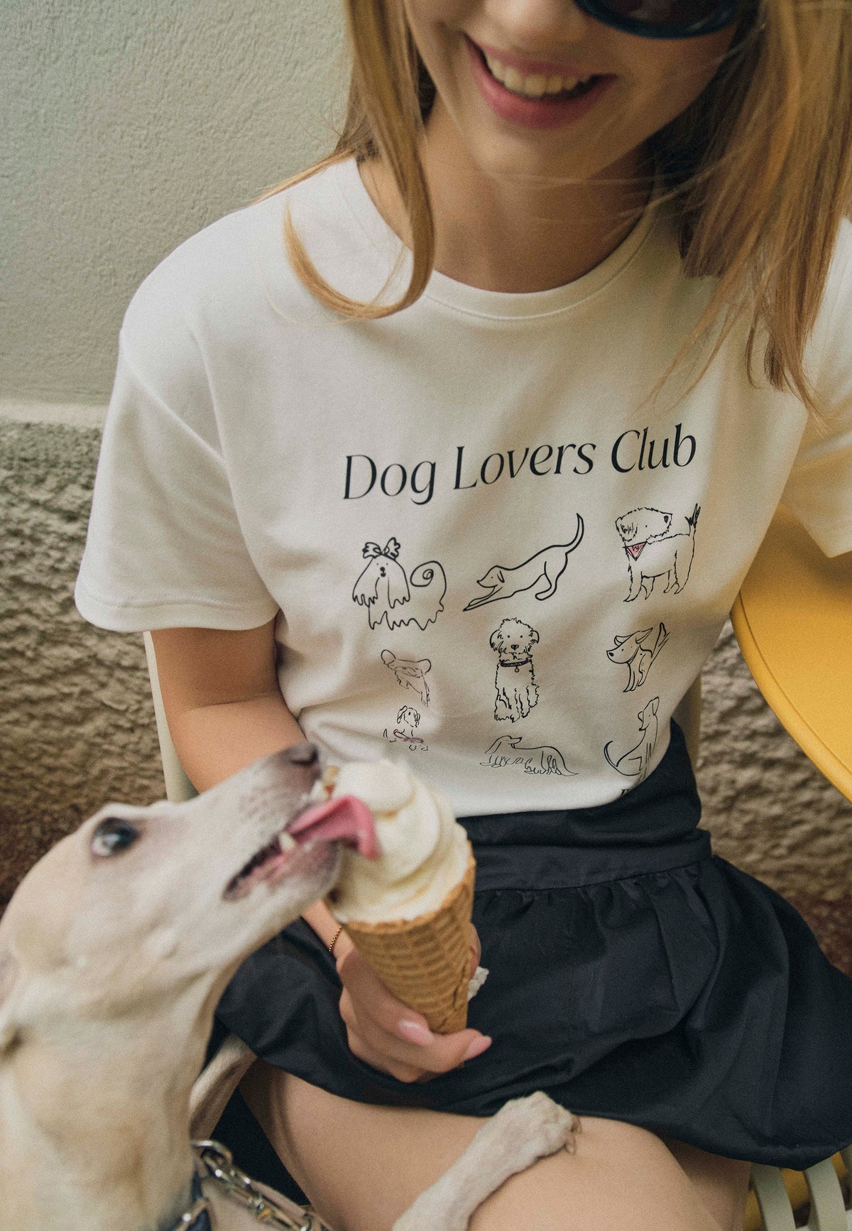 Close-up of a model wearing a T-shirt with drawings of dogs and the words “Dog Lovers Club.” The model is holding an ice cream cone, which a dog is licking.