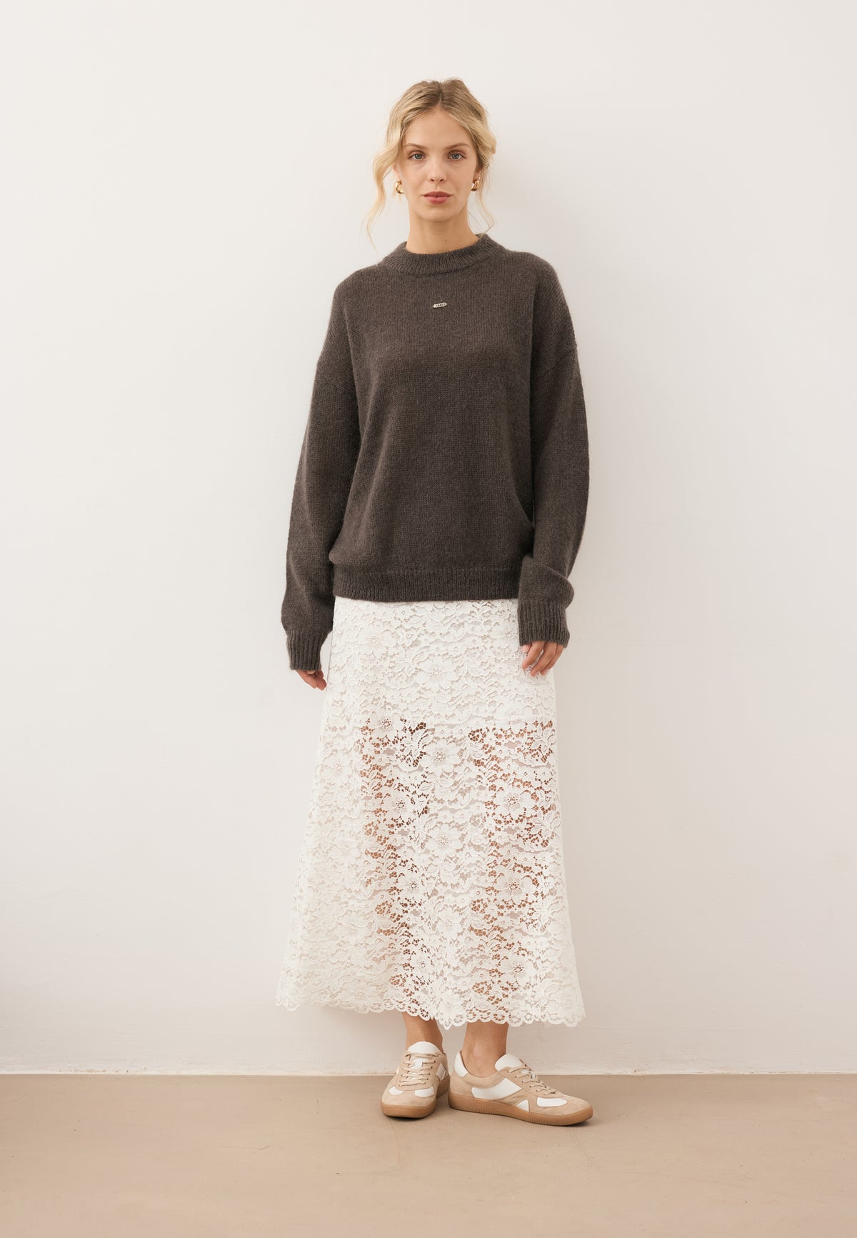 Model posing against a white wall in the brown wool FAROA sweater with front logo and the MILKY lace mini skirt, styled with SODDY sneakers.
