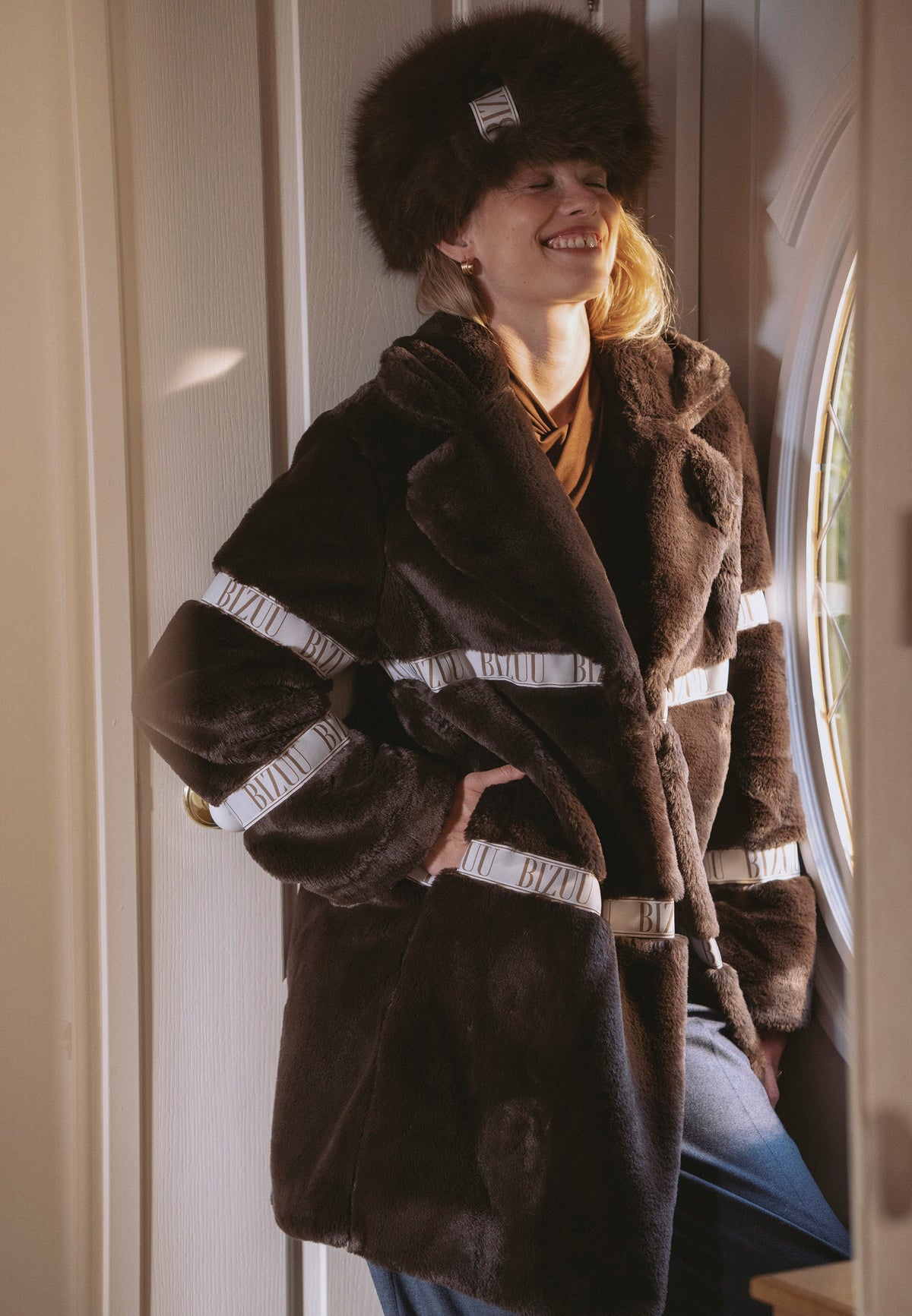 Model leaning against a wall with hands in the pockets of the brown FLORENCE faux fur coat with branded stripes.