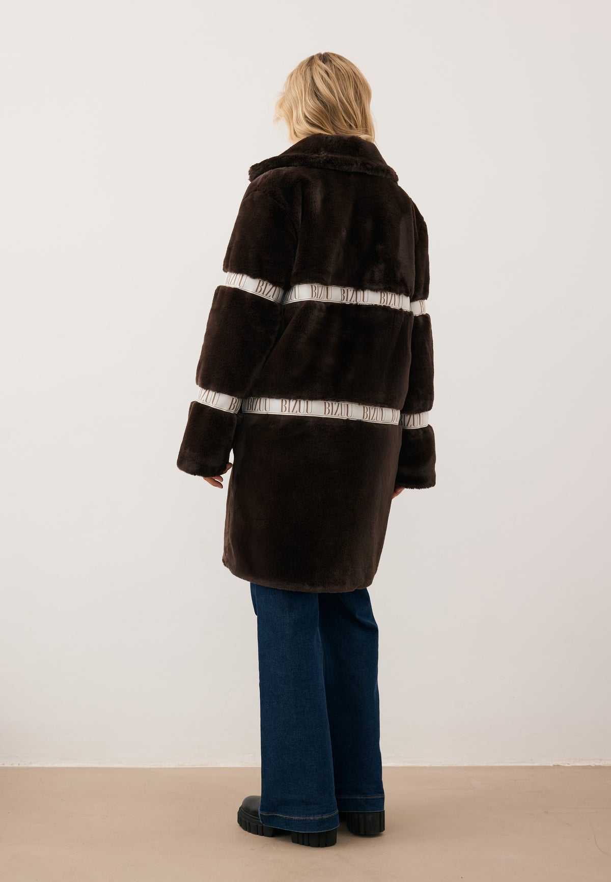Model standing with her back to the camera, wearing the brown FLORENCE faux fur coat with BIZUU logo.